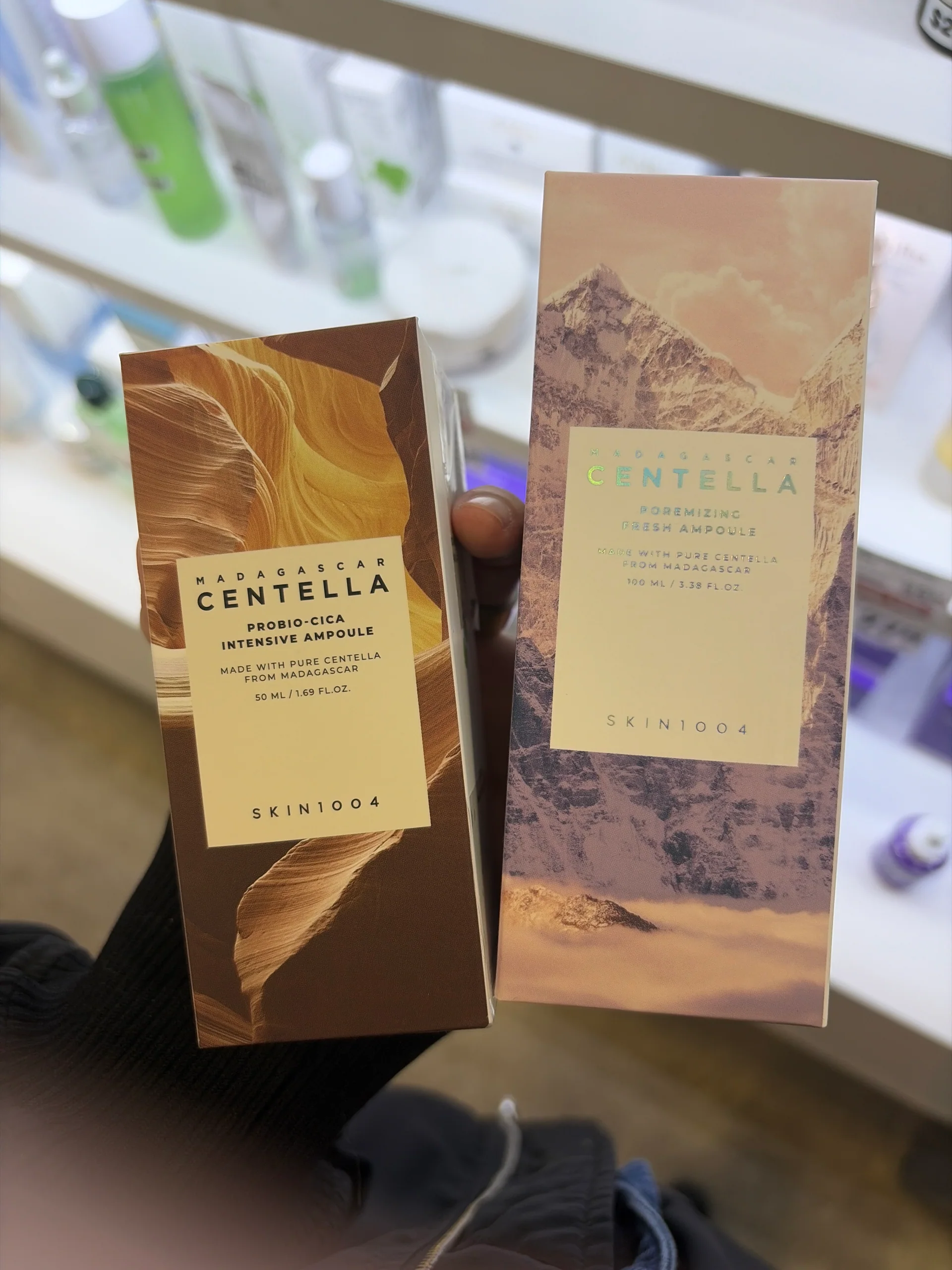 A hand holding SKIN1004 Probio-Cica Intensive Ampoule and Poremizing Fresh Ampoule cartons.
